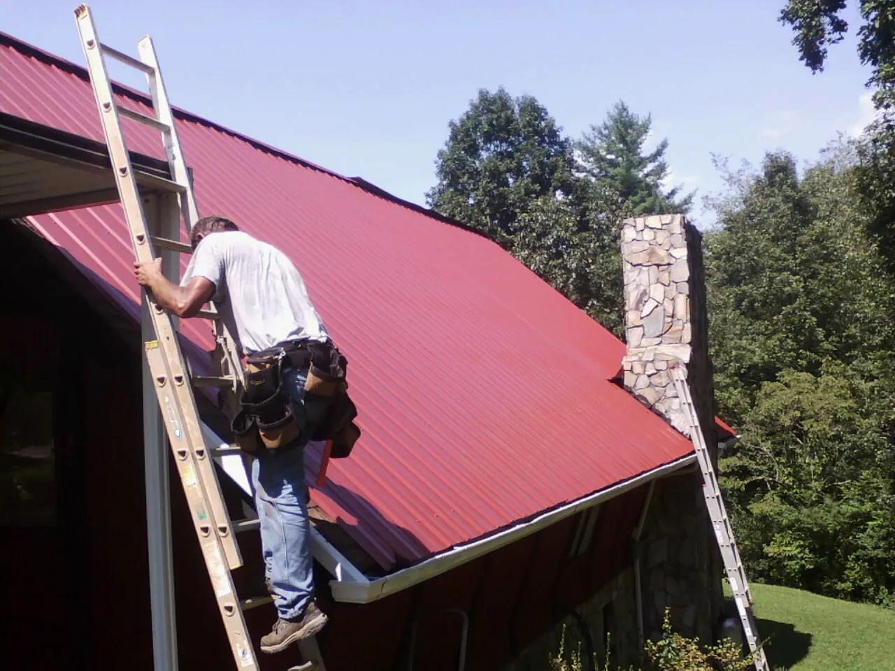 Roofer on a ladder beside a red metal roof and stone chimney — Soffit & Fascia Repair by Roofing Brothers in Yellow Springs